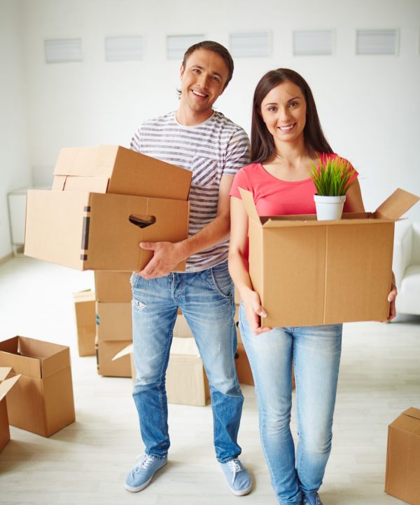Happy young couple with boxes looking at camera in new flat